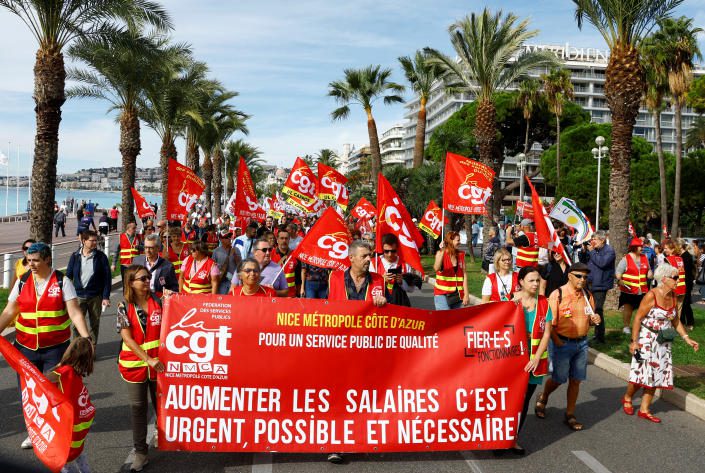 Französische Gewerkschafter gehen eine von Palmen gesäumte Straße entlang und schwenken scharlachrote Fahnen und ein passendes Transparent.